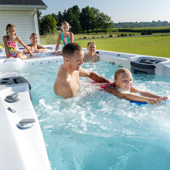 Een vader die zijn dochter op een kickboard helpt om tegen de stroming in te zwemmen in een zwemspa, terwijl de rest van het gezin toekijkt.